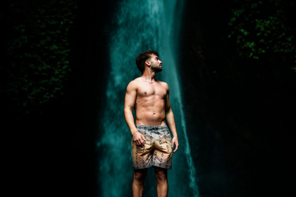 A young man stands confidently before a majestic waterfall in Bali's lush outdoors.