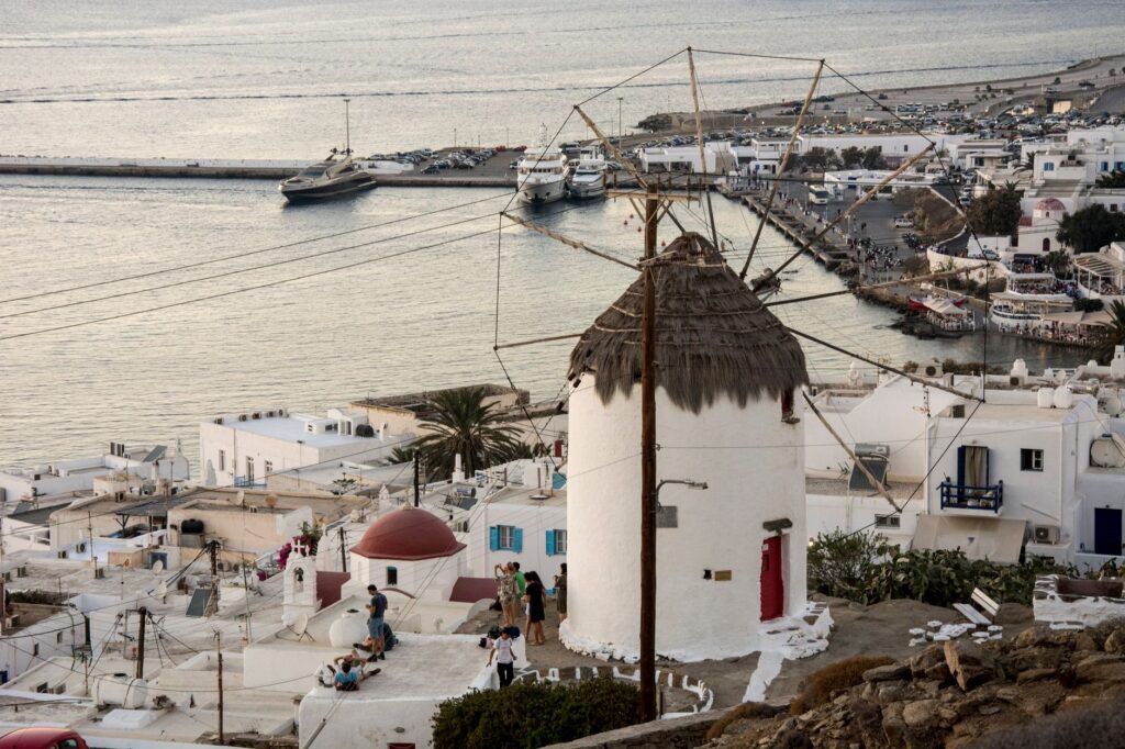Picturesque view of Mykonos windmills, white buildings, and harbor at sunset.