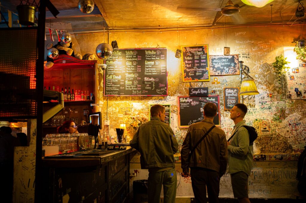 Three people in a Budapest ruin pub, discussing drinks under vibrant lighting and eclectic decor.