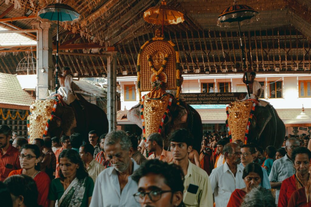 Crowded Indian festival scene featuring ornately decorated elephants under a thatched roof structure.
