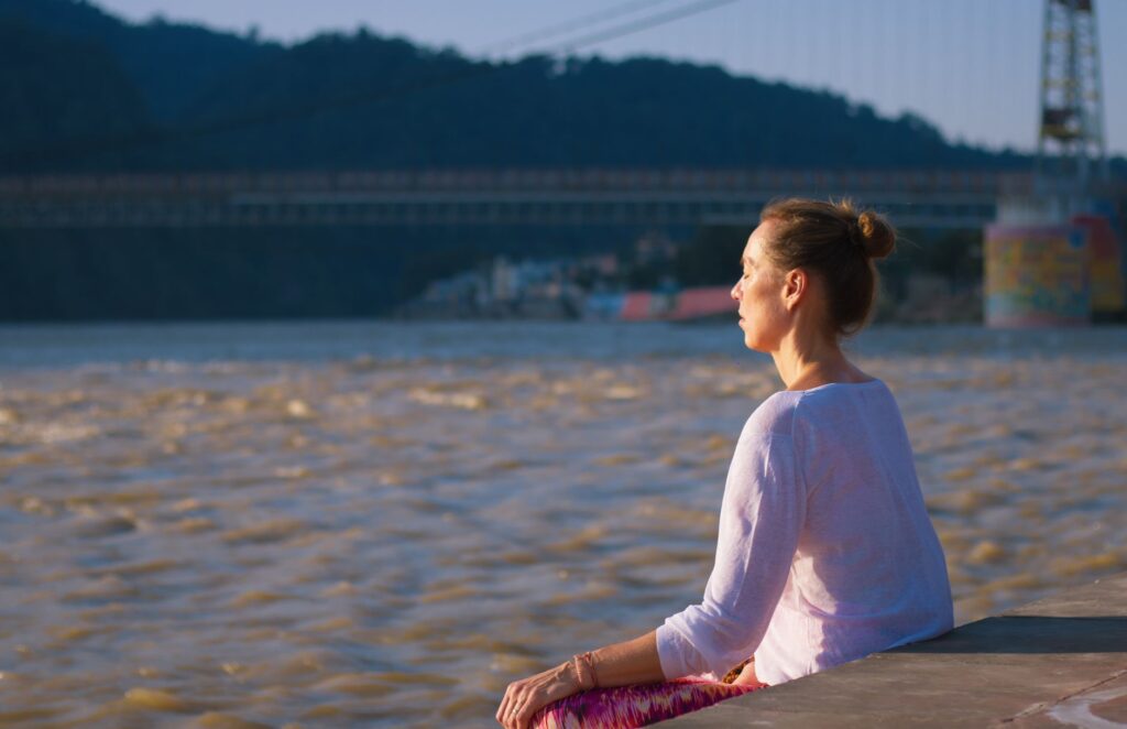 Woman meditating near Ganges River in Rishikesh, India with a bridge in the background.