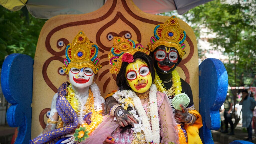 Three children in vibrant traditional costumes at a cultural festival, adorned with colorful face paint and headgear.