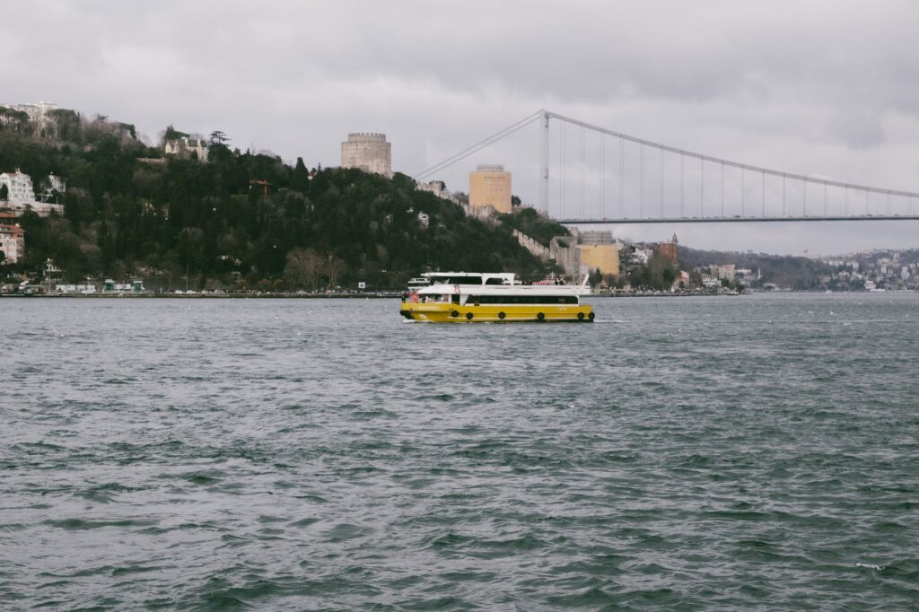 A yellow boat sails on the Bosphorus with the iconic Istanbul bridge in the background.