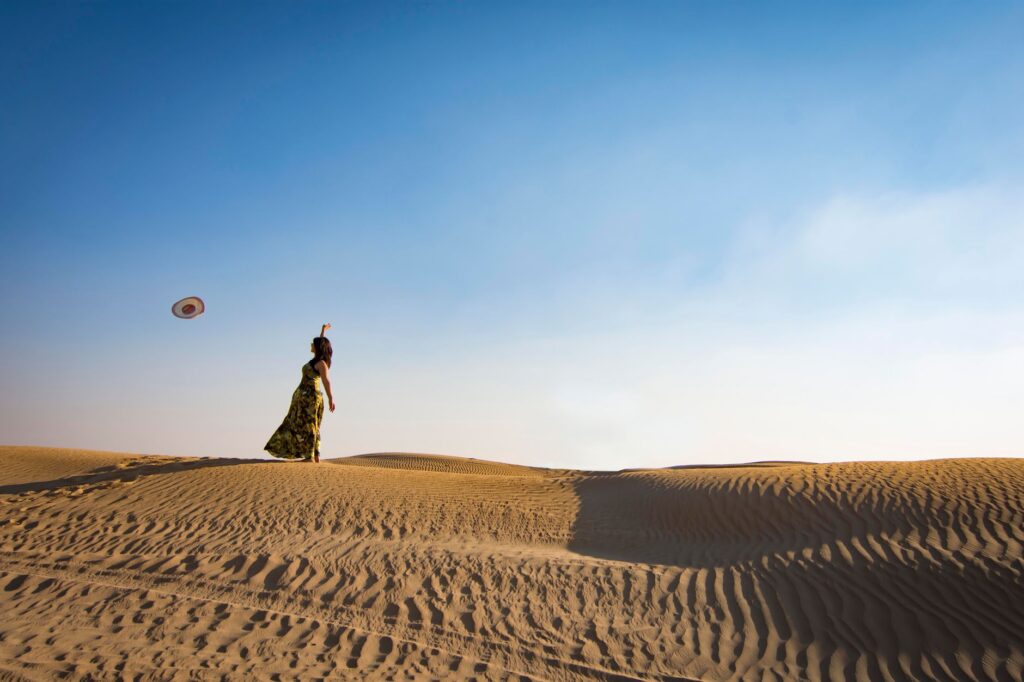 A woman stands alone on sand dunes in Dubai, casting her hat into the air under a clear blue sky.
